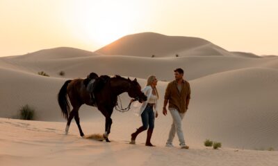 A man and woman walk side by side across Abu Dhabi desert sand dunes at sunset, leading a saddled horse over rippled golden sands.