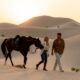 A man and woman walk side by side across Abu Dhabi desert sand dunes at sunset, leading a saddled horse over rippled golden sands.