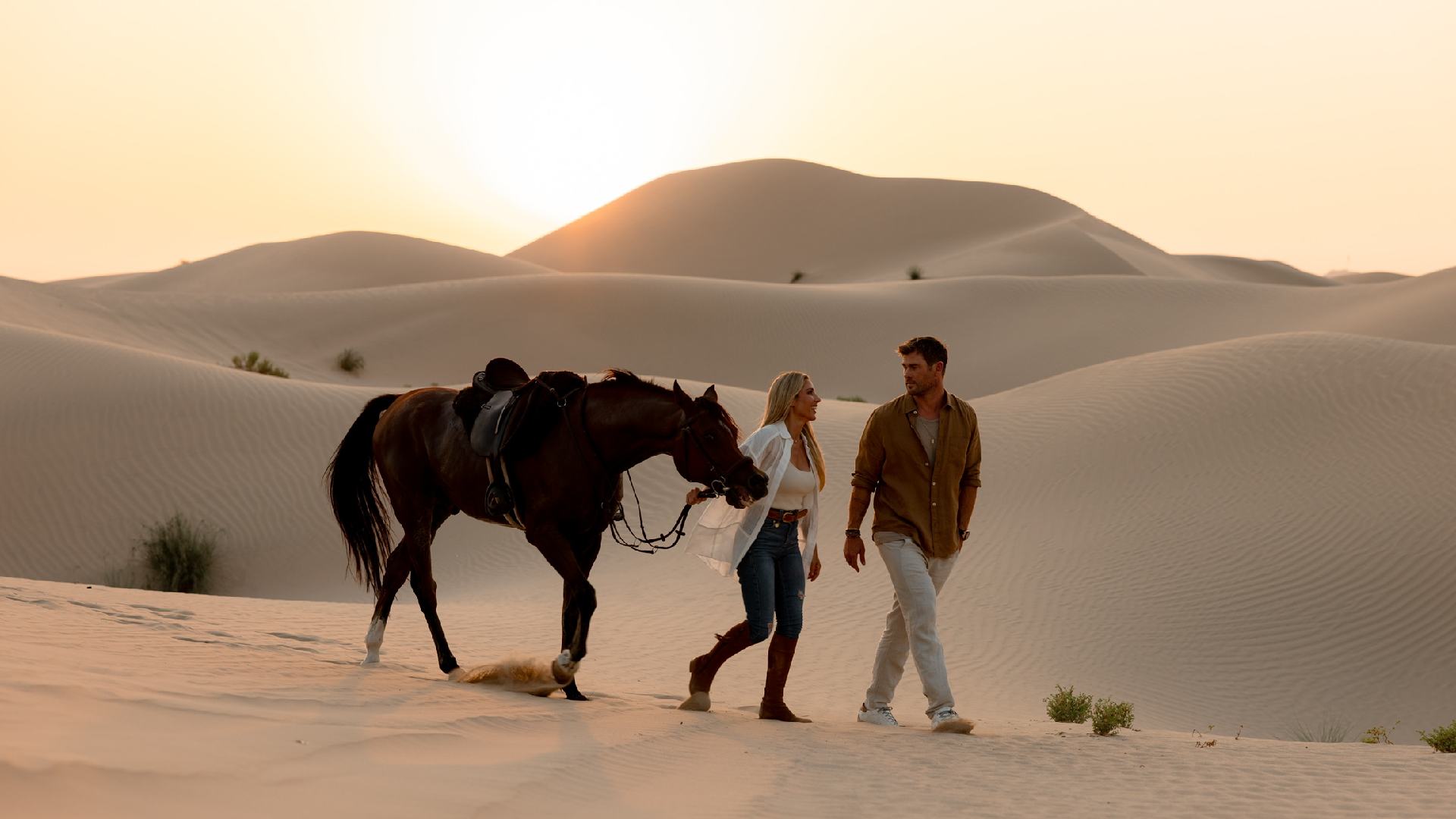 A man and woman walk side by side across Abu Dhabi desert sand dunes at sunset, leading a saddled horse over rippled golden sands.