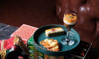 A styled plate of Indian desserts from Rohini, featuring assorted mithai and sweets arranged on a teal platter, including a layered dessert served in a glass, a rectangular mithai slice, and syrup-soaked sweets, displayed on colorful festive boxes against a leather seating backdrop.