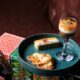 A styled plate of Indian desserts from Rohini, featuring assorted mithai and sweets arranged on a teal platter, including a layered dessert served in a glass, a rectangular mithai slice, and syrup-soaked sweets, displayed on colorful festive boxes against a leather seating backdrop.