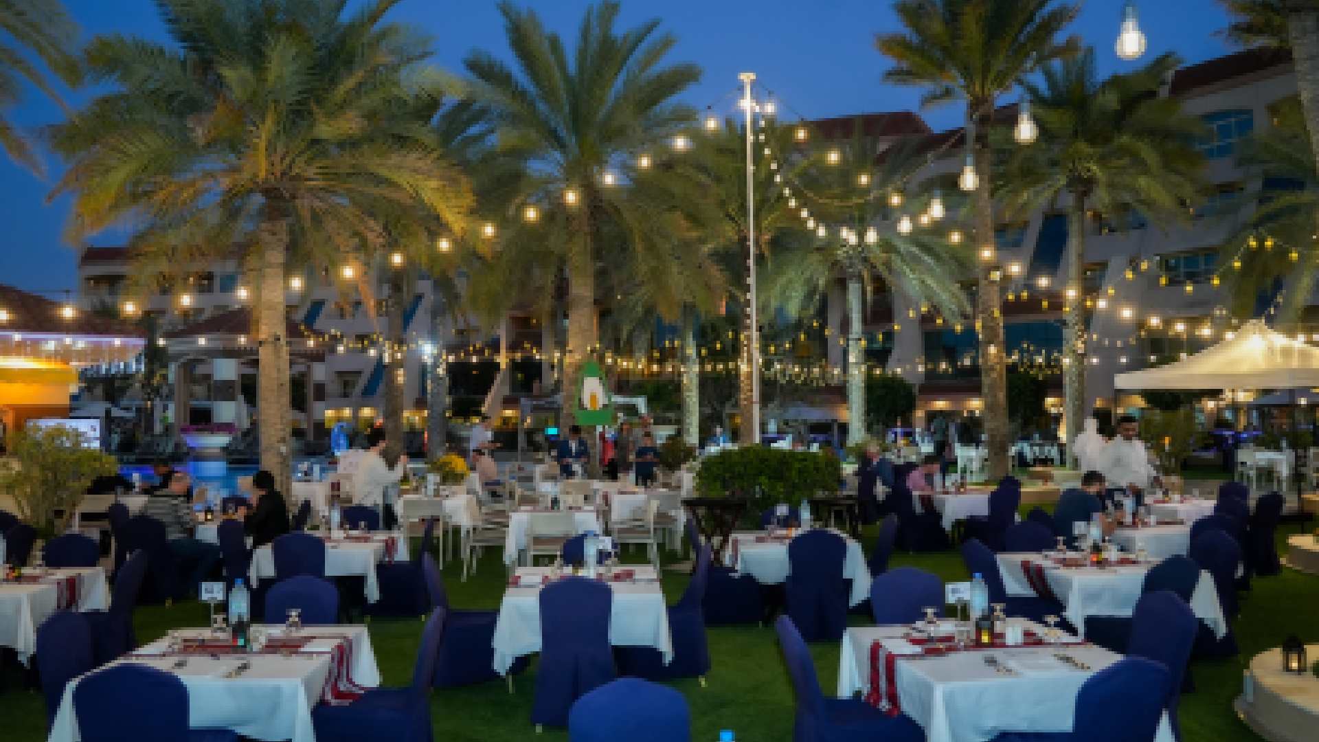 Outdoor Iftar dining setup at AL RAHA BEACH RESORT & SPA featuring beachfront tables under palm trees with warm string lights during evening.