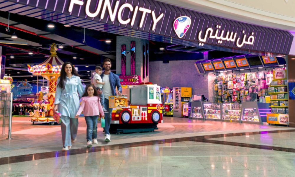 Entrance of Fun City at Ibn Battuta Mall featuring a colorful arcade area with bright signage, toy display counters, and a red-and-white kiddie ride shaped like a fire truck. A family is walking out of the entertainment center, and a carousel is visible inside.