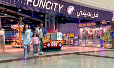 Entrance of Fun City at Ibn Battuta Mall featuring a colorful arcade area with bright signage, toy display counters, and a red-and-white kiddie ride shaped like a fire truck. A family is walking out of the entertainment center, and a carousel is visible inside.