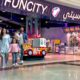 Entrance of Fun City at Ibn Battuta Mall featuring a colorful arcade area with bright signage, toy display counters, and a red-and-white kiddie ride shaped like a fire truck. A family is walking out of the entertainment center, and a carousel is visible inside.