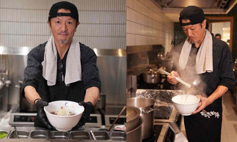 Chef standing in a professional kitchen holding a bowl of ramen with both hands, wearing black gloves, a dark shirt, and a towel draped around his neck, with stainless‑steel counters and cooking equipment in the background.