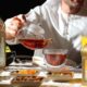 Man pours hot Arabic tea from glass teapot into a clear cup on a Ramadan table with lanterns and dates at Hilton Garden Inn Dubai.