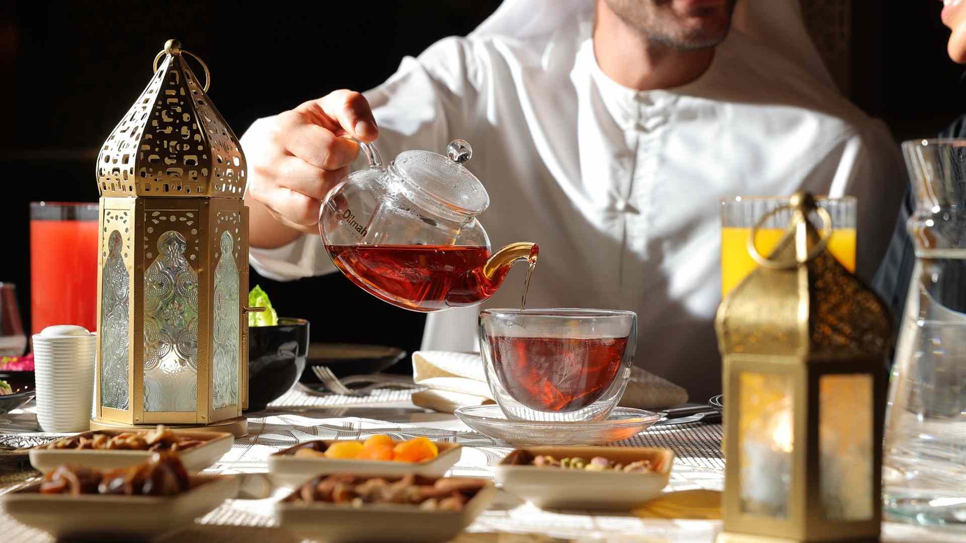 Man pours hot Arabic tea from glass teapot into a clear cup on a Ramadan table with lanterns and dates at Hilton Garden Inn Dubai.