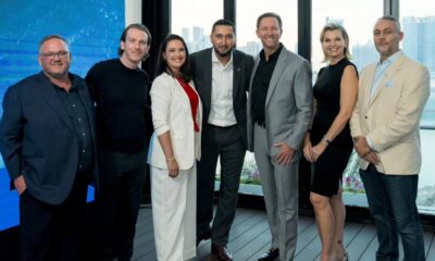 Group of six people standing together indoors near large windows with a city skyline and waterfront view in the background, next to a blue digital display screen showing the Hashgraph Ventures logo