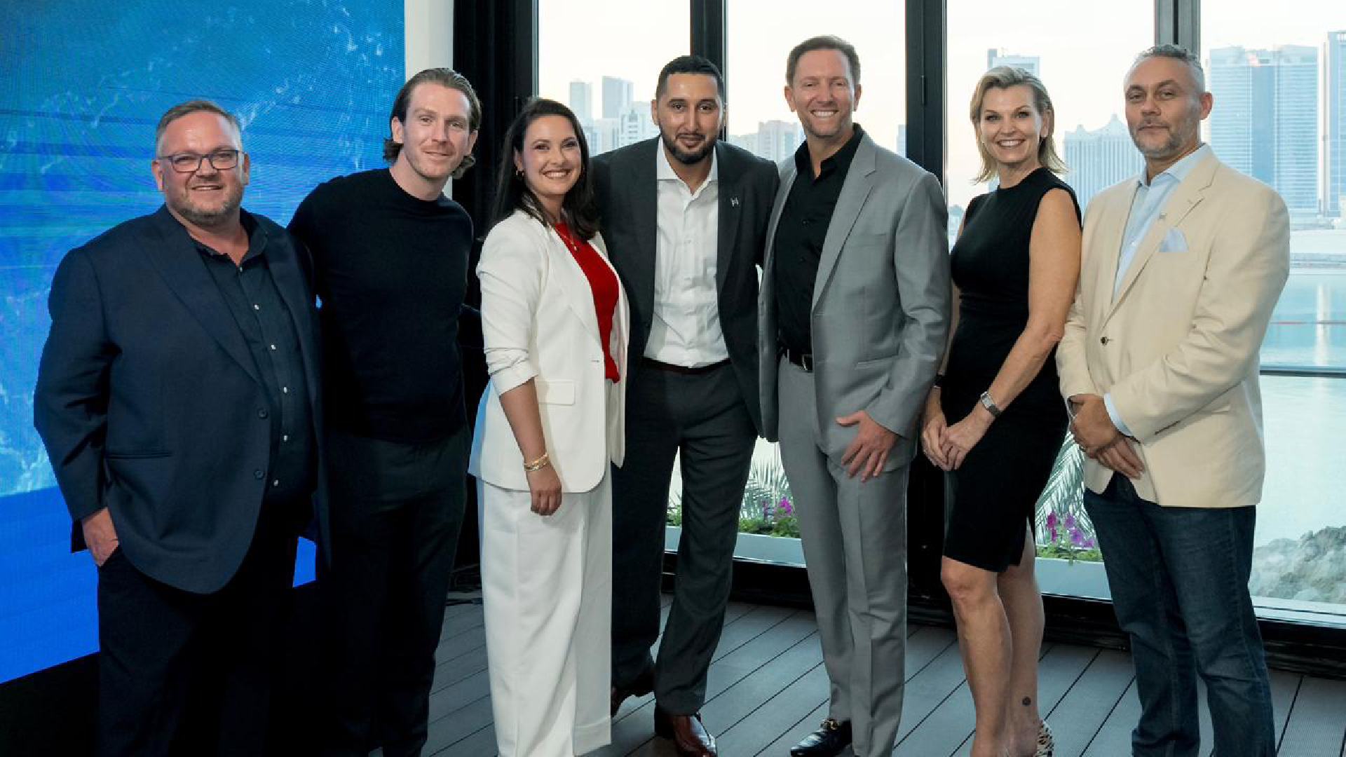 Group of six people standing together indoors near large windows with a city skyline and waterfront view in the background, next to a blue digital display screen showing the Hashgraph Ventures logo