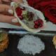 Hand holding an oyster garnished with red berries and microgreens above a black serving plate featuring salmon tartare on the left, coarse sea salt in the center, and a tempura shrimp on the right, with red rose petals and a bouquet of roses in the background.