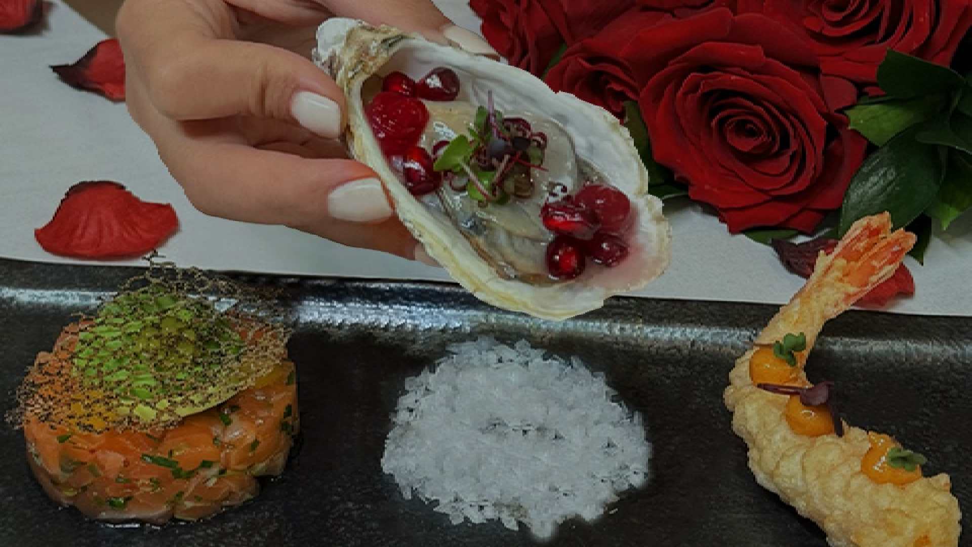Hand holding an oyster garnished with red berries and microgreens above a black serving plate featuring salmon tartare on the left, coarse sea salt in the center, and a tempura shrimp on the right, with red rose petals and a bouquet of roses in the background.