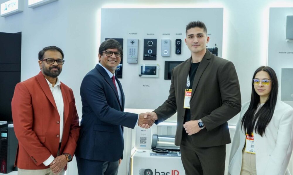 A ScreenCheck representative and a BAS-IP representative shake hands in front of a display showcasing BAS-IP intercom and security devices, with two additional team members standing beside them at an exhibition booth.