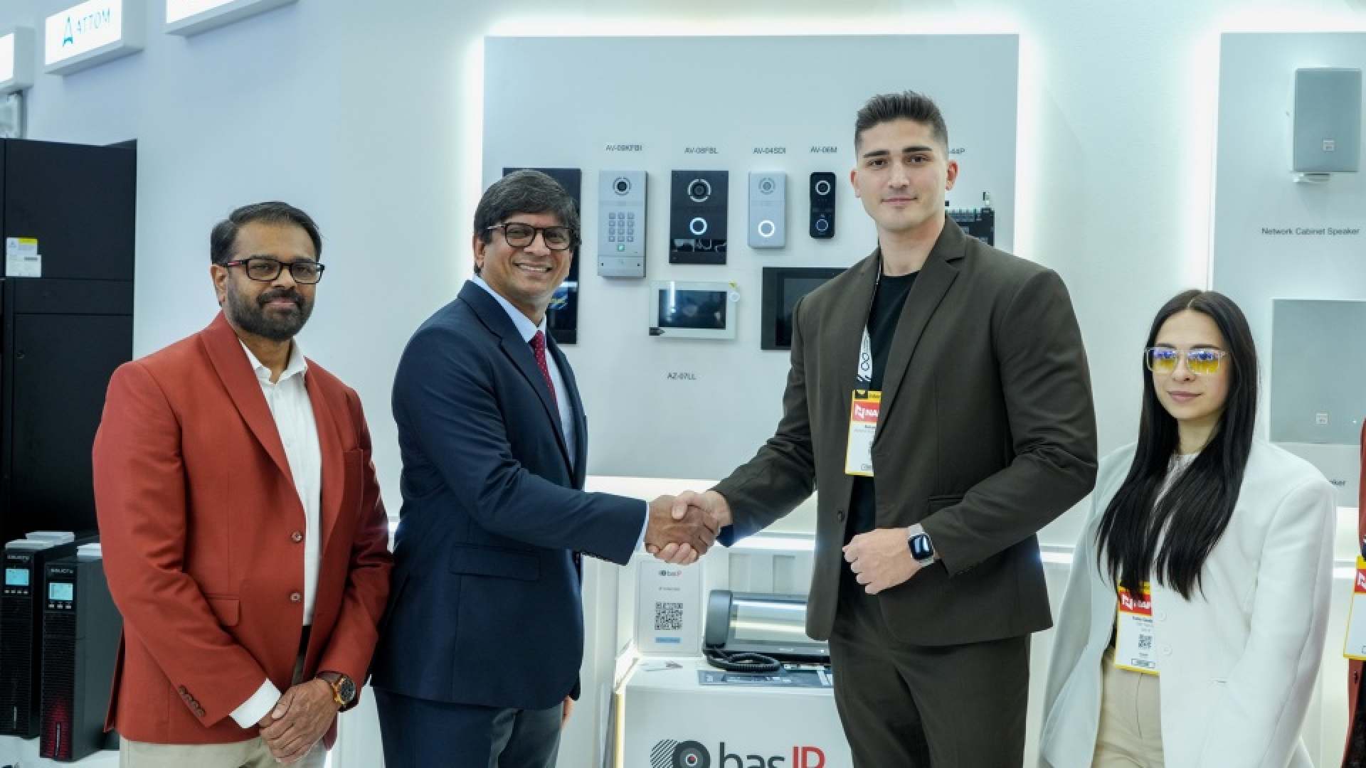 A ScreenCheck representative and a BAS-IP representative shake hands in front of a display showcasing BAS-IP intercom and security devices, with two additional team members standing beside them at an exhibition booth.