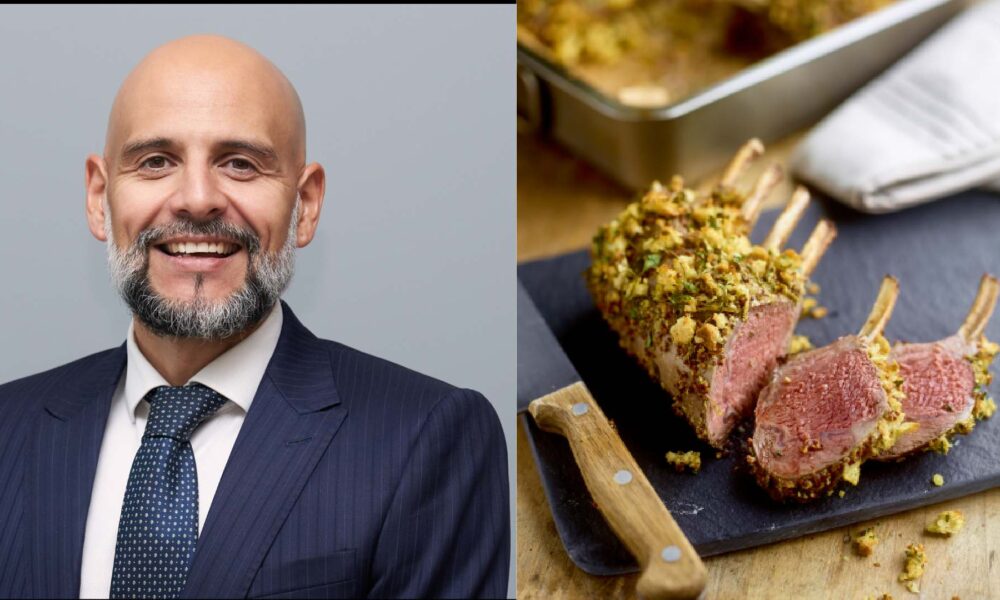 A man wearing a dark pinstripe suit, white shirt and navy tie standing for a professional portrait next to a sliced herb‑crusted rack of lamb served on a cutting board with a wooden‑handled knife.