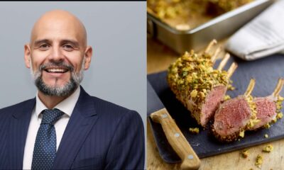 A man wearing a dark pinstripe suit, white shirt and navy tie standing for a professional portrait next to a sliced herb‑crusted rack of lamb served on a cutting board with a wooden‑handled knife.