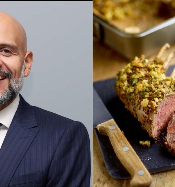 A man wearing a dark pinstripe suit, white shirt and navy tie standing for a professional portrait next to a sliced herb‑crusted rack of lamb served on a cutting board with a wooden‑handled knife.