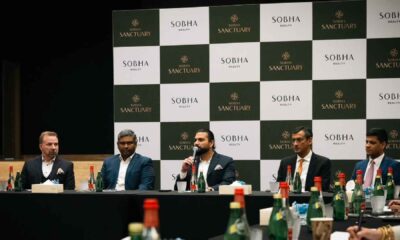 A Sobha Realty press conference panel with company representatives seated at a table, speaking into microphones, with Sobha Sanctuary and Sobha Realty branded backdrop behind them and water bottles arranged on the table.