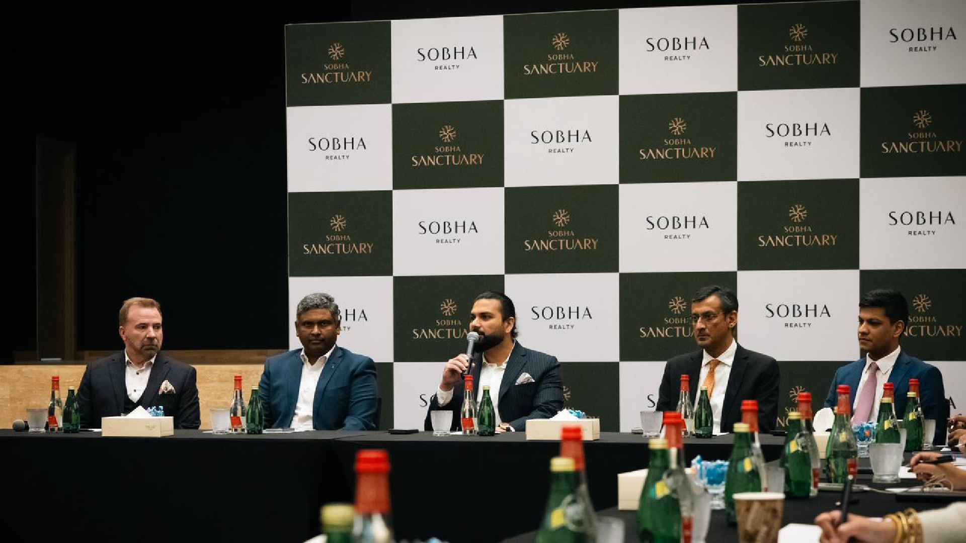 A Sobha Realty press conference panel with company representatives seated at a table, speaking into microphones, with Sobha Sanctuary and Sobha Realty branded backdrop behind them and water bottles arranged on the table.