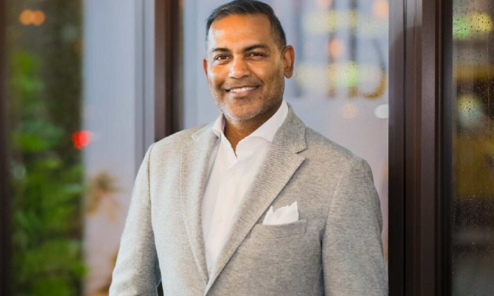 A person wearing a light grey blazer, white dress shirt, and pocket square stands indoors near large glass windows with soft ambient lighting and greenery visible in the background. Photo provided by Sun Siyam.