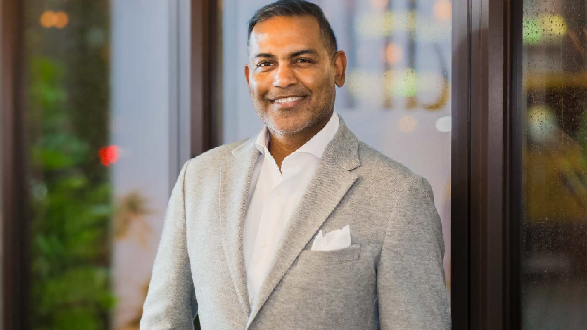 A person wearing a light grey blazer, white dress shirt, and pocket square stands indoors near large glass windows with soft ambient lighting and greenery visible in the background. Photo provided by Sun Siyam.