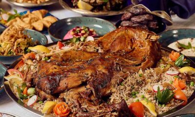 A large platter of slow-roasted lamb served over spiced rice and garnished with vegetables, surrounded by assorted Iftar dishes including salads, dates, samosas, and side plates on a buffet table at Spice Island.