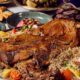 A large platter of slow-roasted lamb served over spiced rice and garnished with vegetables, surrounded by assorted Iftar dishes including salads, dates, samosas, and side plates on a buffet table at Spice Island.
