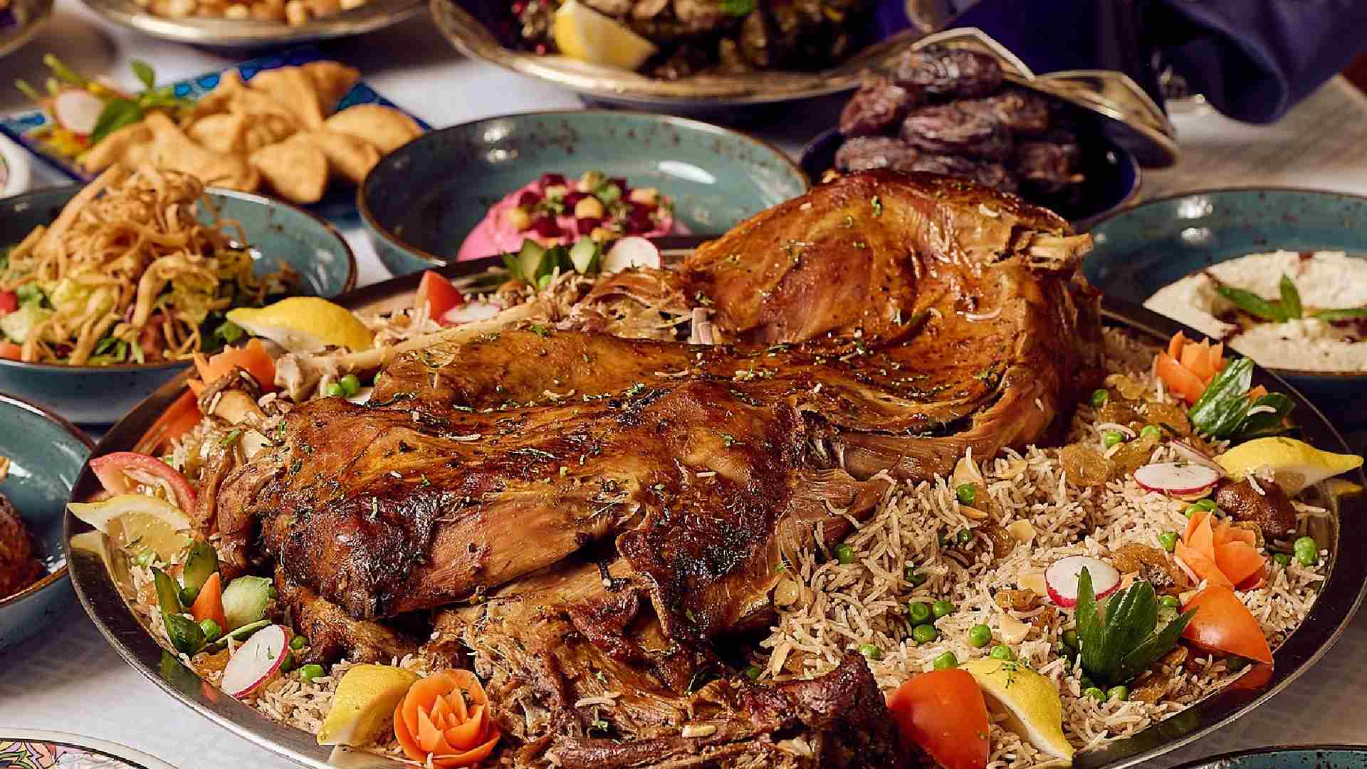 A large platter of slow-roasted lamb served over spiced rice and garnished with vegetables, surrounded by assorted Iftar dishes including salads, dates, samosas, and side plates on a buffet table at Spice Island.