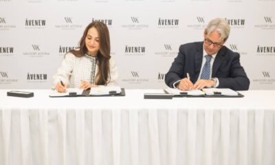 A signing ceremony where two individuals seated at a table are signing partnership documents in front of a backdrop displaying the AVENEW Development and Waldorf Astoria logos.
