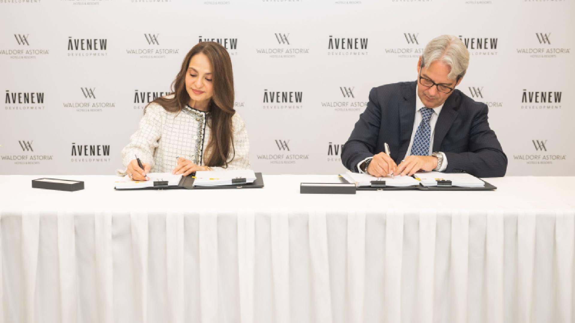 A signing ceremony where two individuals seated at a table are signing partnership documents in front of a backdrop displaying the AVENEW Development and Waldorf Astoria logos.