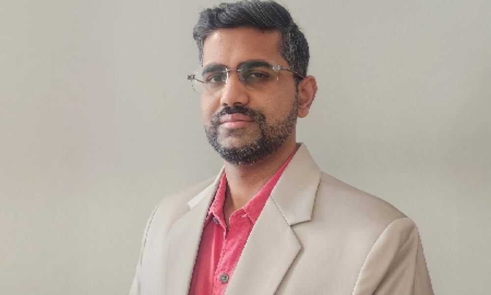 Person wearing a beige suit jacket over a red collared shirt, standing against a plain light-colored background.