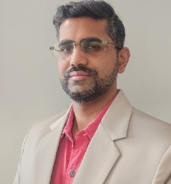 Person wearing a beige suit jacket over a red collared shirt, standing against a plain light-colored background.