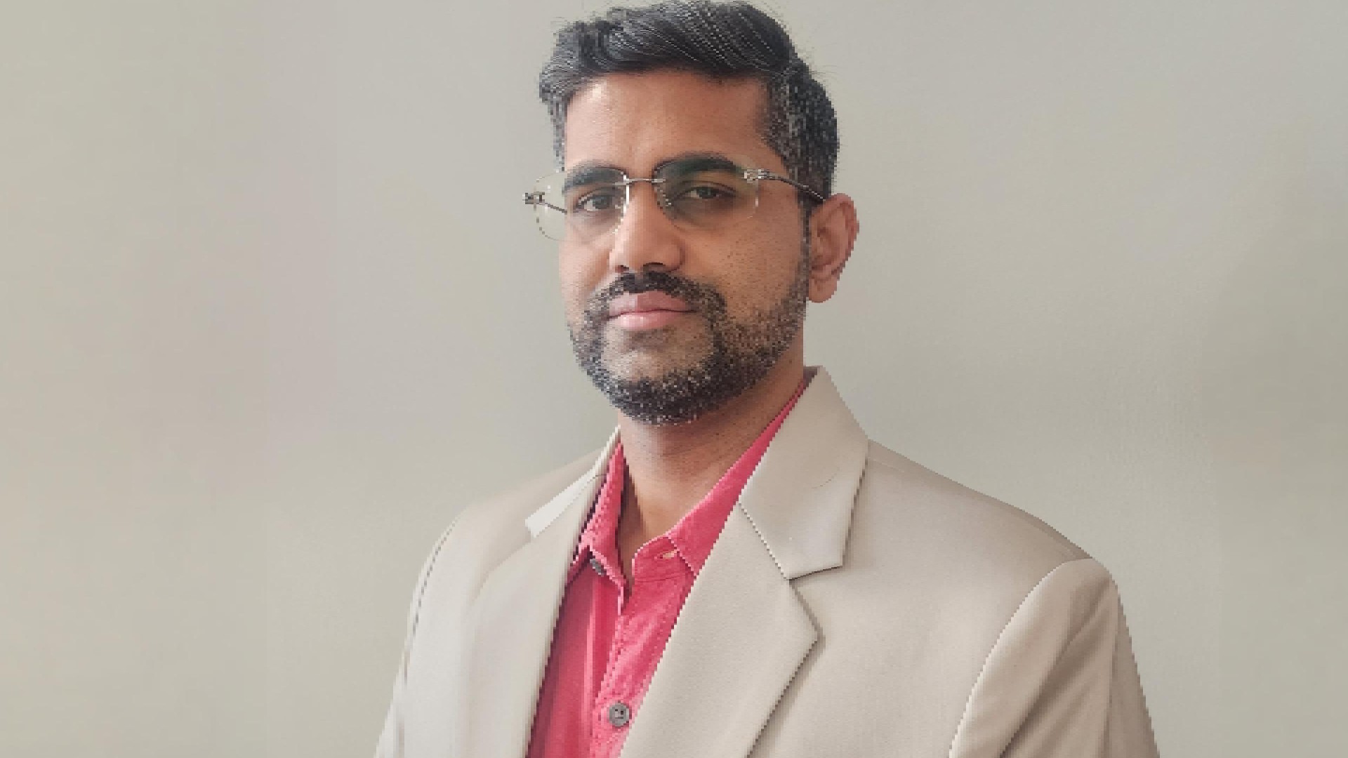 Person wearing a beige suit jacket over a red collared shirt, standing against a plain light-colored background.