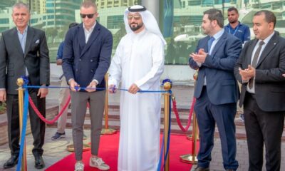 Ribbon-cutting ceremony on a red carpet at an outdoor venue with five formally dressed individuals standing in front of gold stanchions and blue ribbon, surrounded by modern high-rise buildings and palm trees in the background.