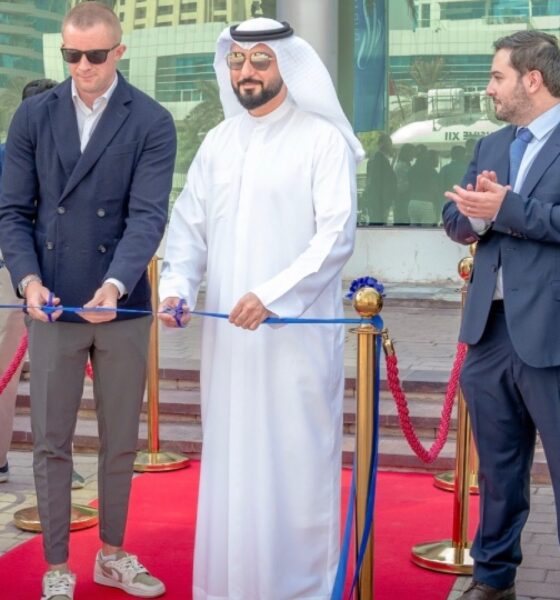 Ribbon-cutting ceremony on a red carpet at an outdoor venue with five formally dressed individuals standing in front of gold stanchions and blue ribbon, surrounded by modern high-rise buildings and palm trees in the background.