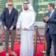 Ribbon-cutting ceremony on a red carpet at an outdoor venue with five formally dressed individuals standing in front of gold stanchions and blue ribbon, surrounded by modern high-rise buildings and palm trees in the background.