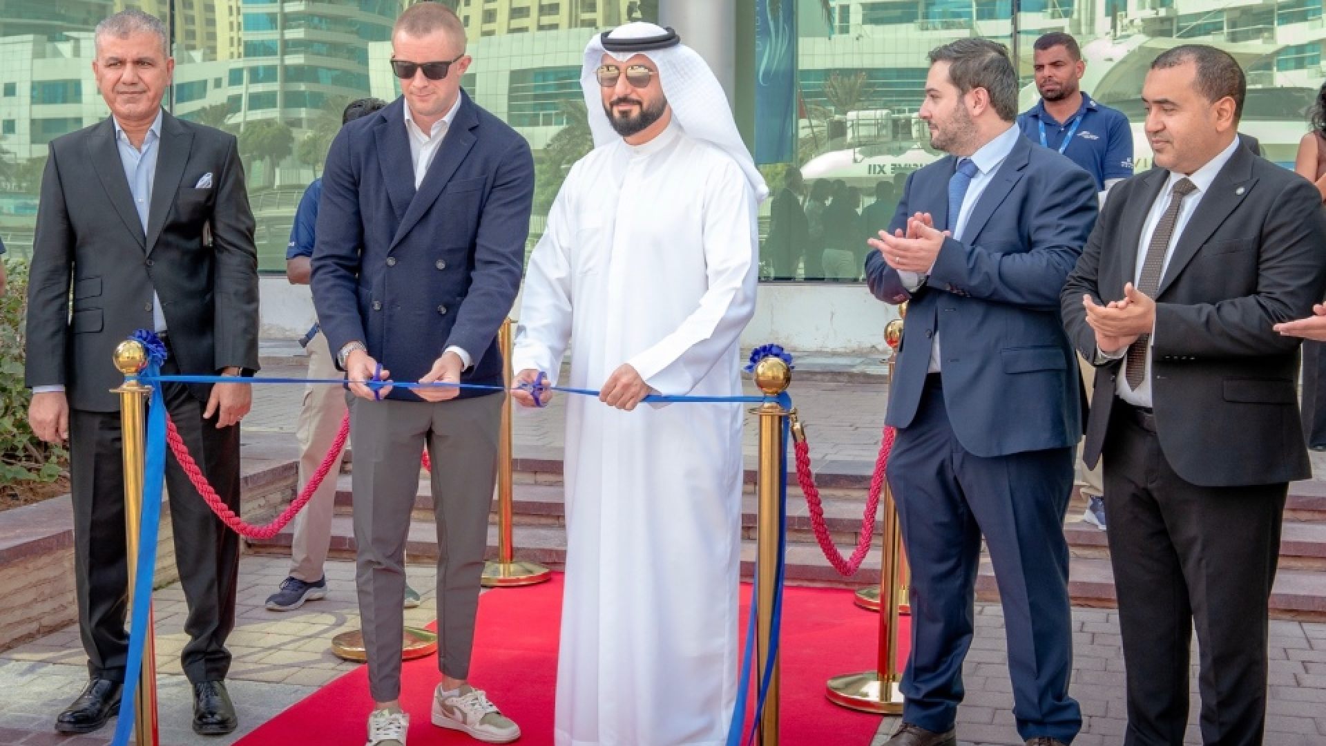 Ribbon-cutting ceremony on a red carpet at an outdoor venue with five formally dressed individuals standing in front of gold stanchions and blue ribbon, surrounded by modern high-rise buildings and palm trees in the background.