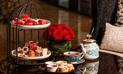 Two tier afternoon tea stand with assorted pastries, teacups, teapot, and red roses on a glass table at Al Jaddaf Rotana.