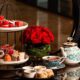Two tier afternoon tea stand with assorted pastries, teacups, teapot, and red roses on a glass table at Al Jaddaf Rotana.