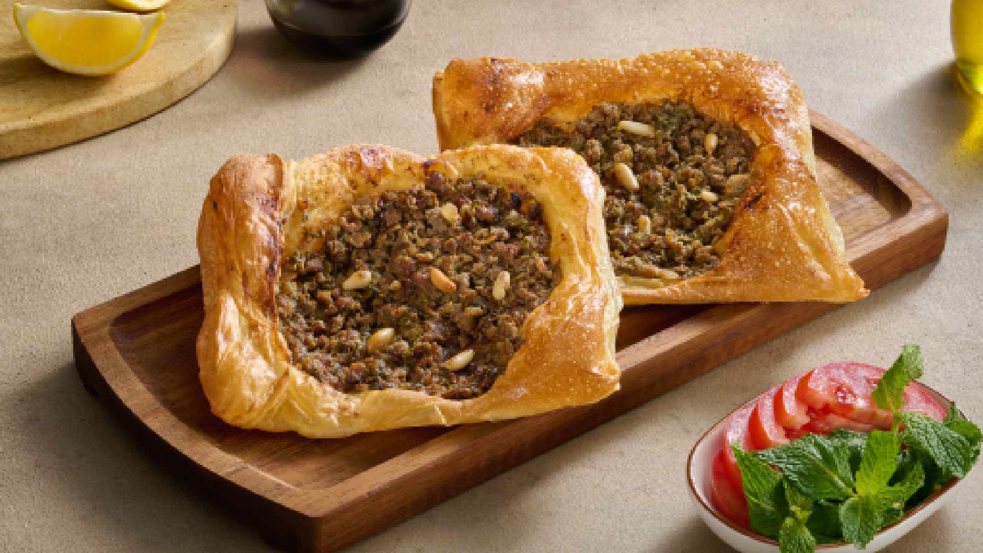 Open Lebanese lahme baajine pastries filled with spiced minced meat, served on a wooden board with lemon, olive oil and fresh salad at AL HALLAB.