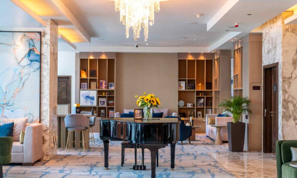 A spacious, modern lounge at voco Bonnington featuring a black grand piano with a vase of sunflowers on top, surrounded by plush seating, marble accents, built‑in bookshelf displays, soft lighting, and a large crystal chandelier overhead.