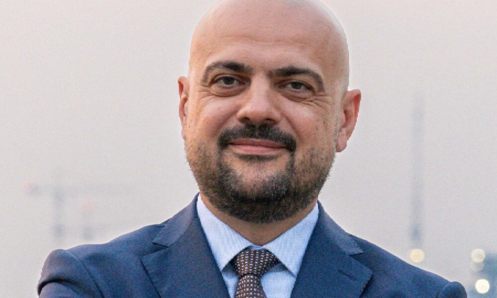 A person in a formal navy suit stands with arms crossed near a waterfront, with city lights and construction cranes visible in the background, related to the RAK property market.