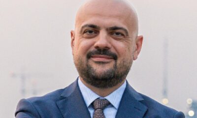 A person in a formal navy suit stands with arms crossed near a waterfront, with city lights and construction cranes visible in the background, related to the RAK property market.