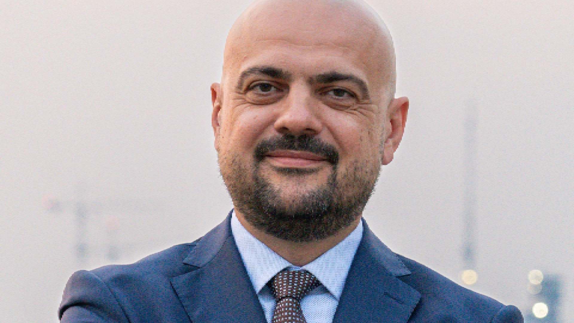 A person in a formal navy suit stands with arms crossed near a waterfront, with city lights and construction cranes visible in the background, related to the RAK property market.