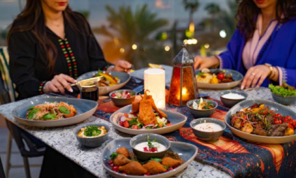 Outdoor Ramadan dining setup at Hilton Abu Dhabi featuring a table filled with Middle Eastern dishes, including grilled meats, mezze, salads, and pastries, with two guests seated and enjoying the meal in a warm, lantern‑lit setting overlooking palm trees and evening views.