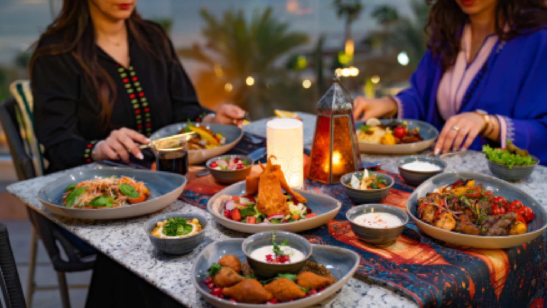 Outdoor Ramadan dining setup at Hilton Abu Dhabi featuring a table filled with Middle Eastern dishes, including grilled meats, mezze, salads, and pastries, with two guests seated and enjoying the meal in a warm, lantern‑lit setting overlooking palm trees and evening views.