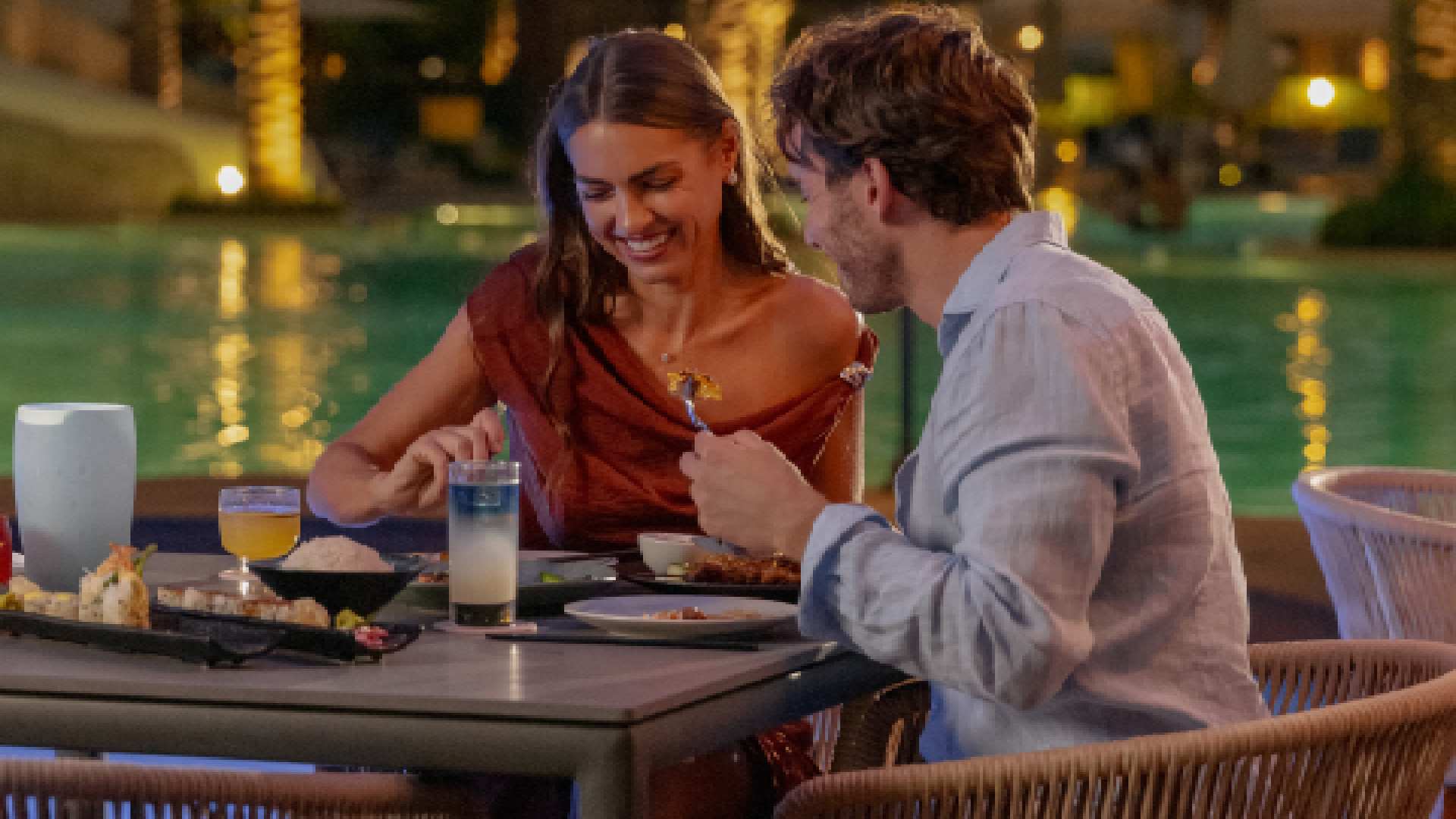 A couple enjoying an intimate outdoor dinner beside a softly lit pool at ONE&ONLY ROYAL MIRAGE, with plated dishes and drinks arranged on the table in a romantic evening setting