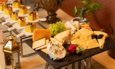 Cheese platter at Pincode featuring assorted cheeses, green grapes, crackers, olives, and strawberries on a black slate, with plated Indian desserts and a decorative lantern in the background.