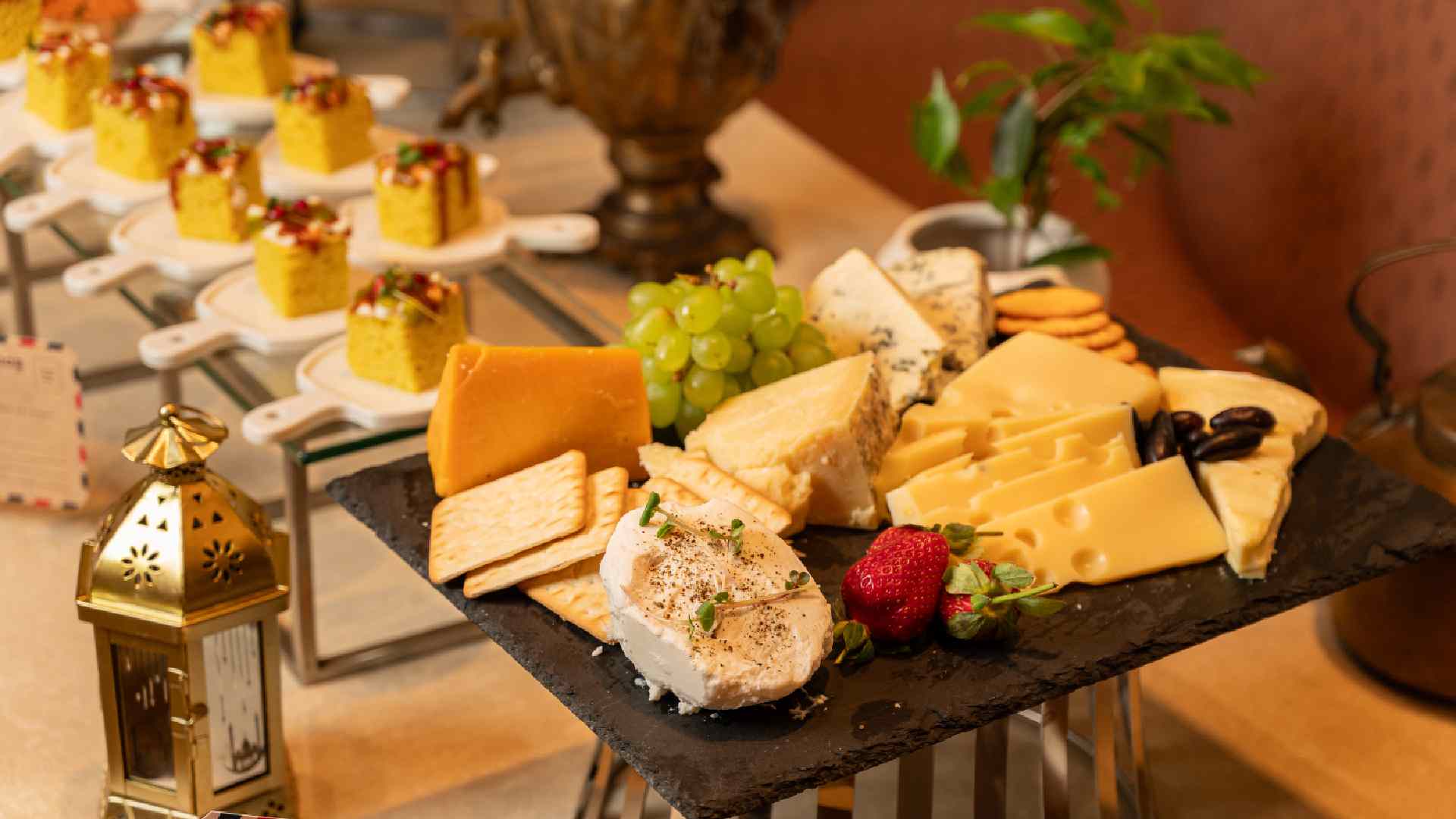 Cheese platter at Pincode featuring assorted cheeses, green grapes, crackers, olives, and strawberries on a black slate, with plated Indian desserts and a decorative lantern in the background.