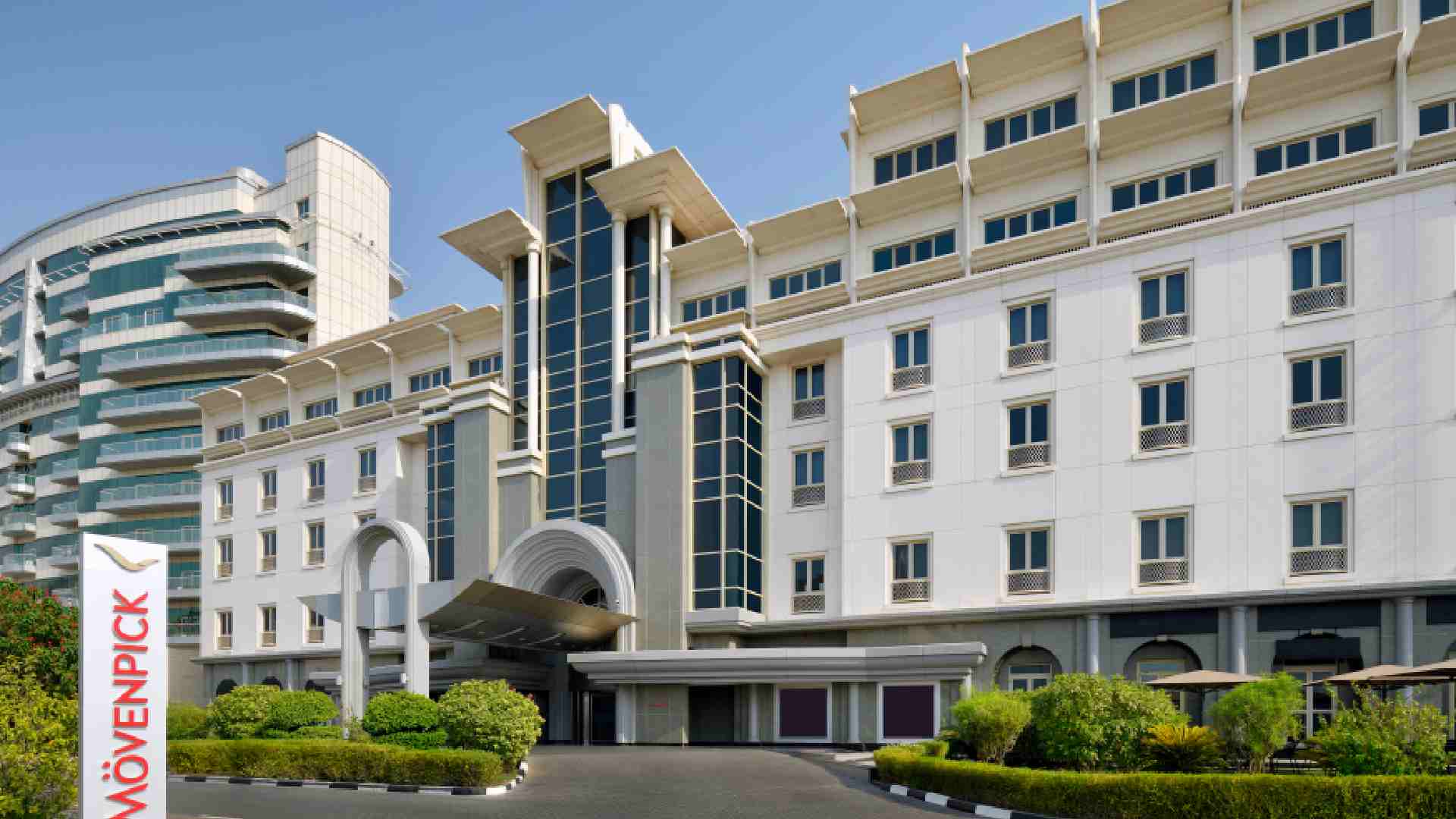 Exterior view of MÖVENPICK HOTEL APARTMENTS Bur Dubai showing the main entrance, white facade, arched driveway, and landscaped front area under clear daylight